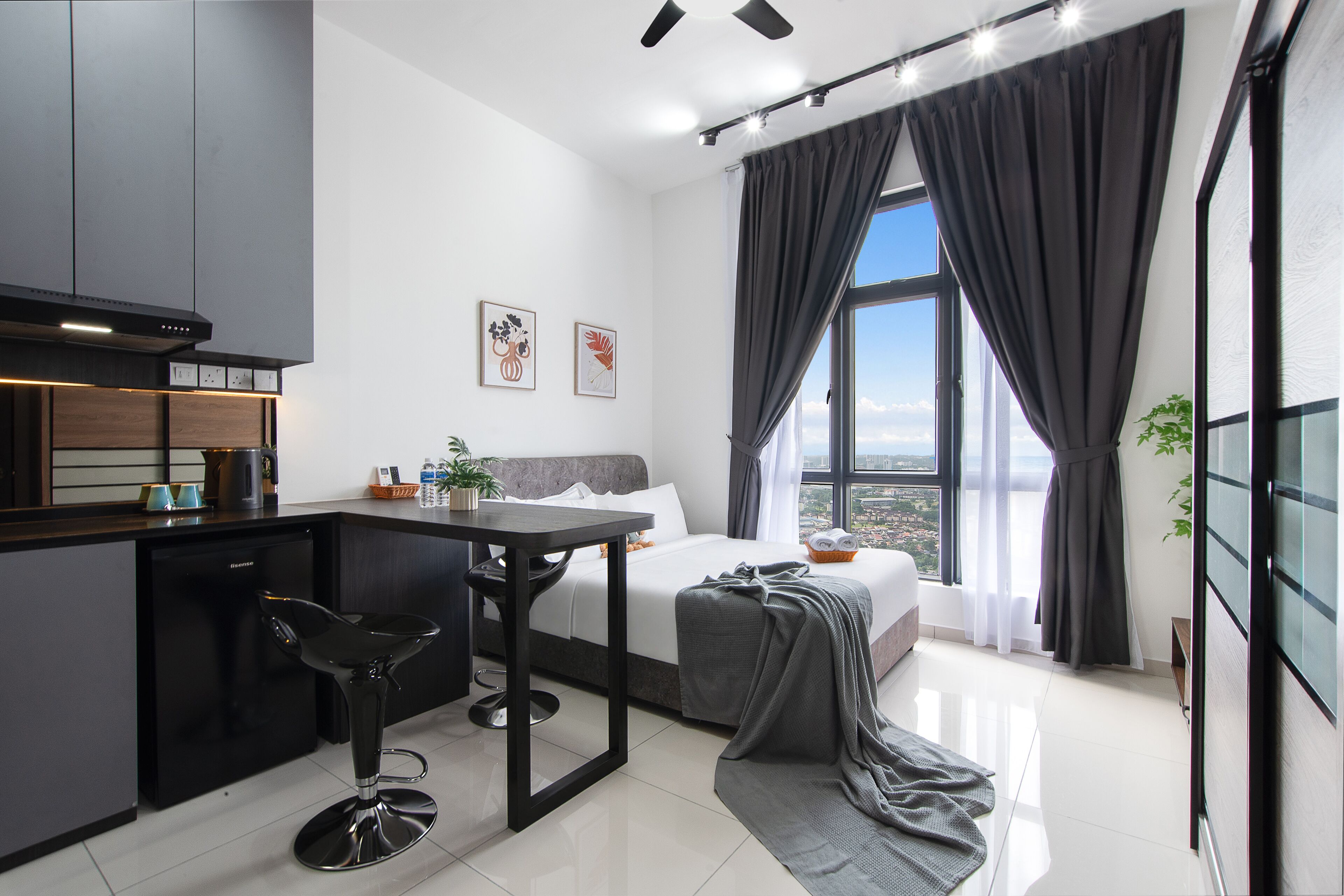 A modern hotel room with a kitchenette, a bed with a view, and a large window with curtains.
