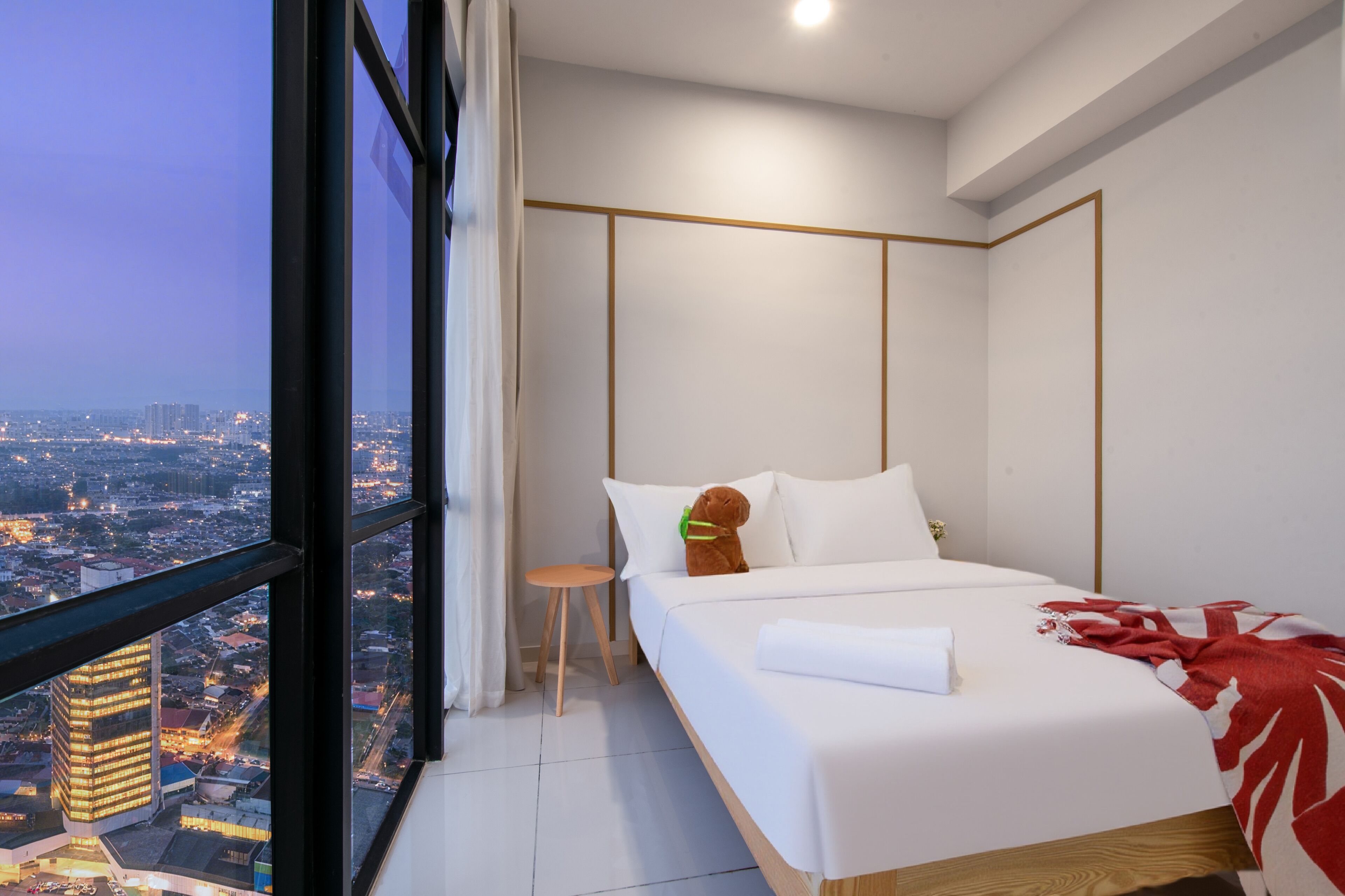 A modern hotel room with a large window, a bed, a small stool, and a view of the cityscape.