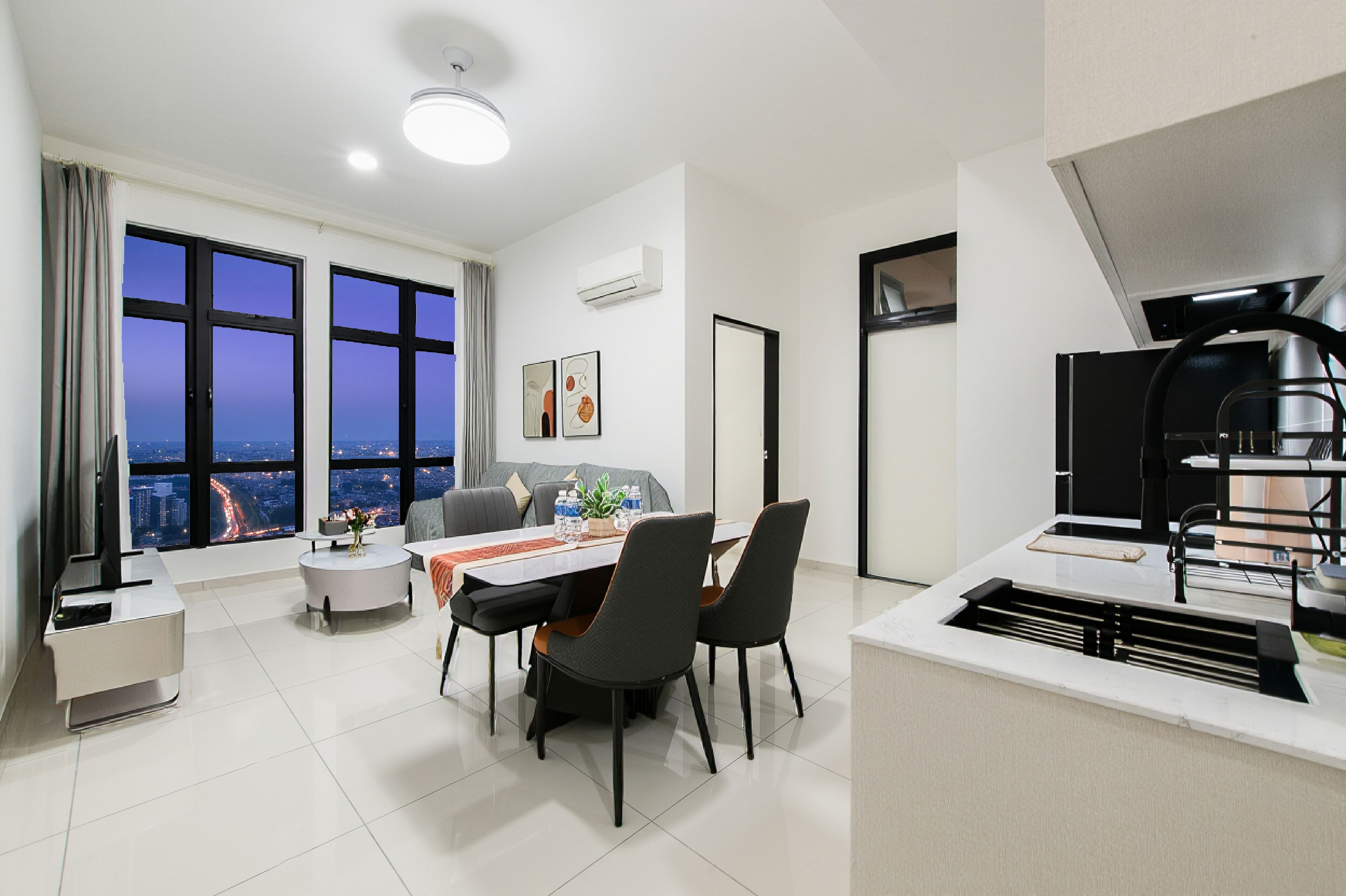 A modern living room with a dining area, large windows with a city view, and a kitchen with a black range hood.