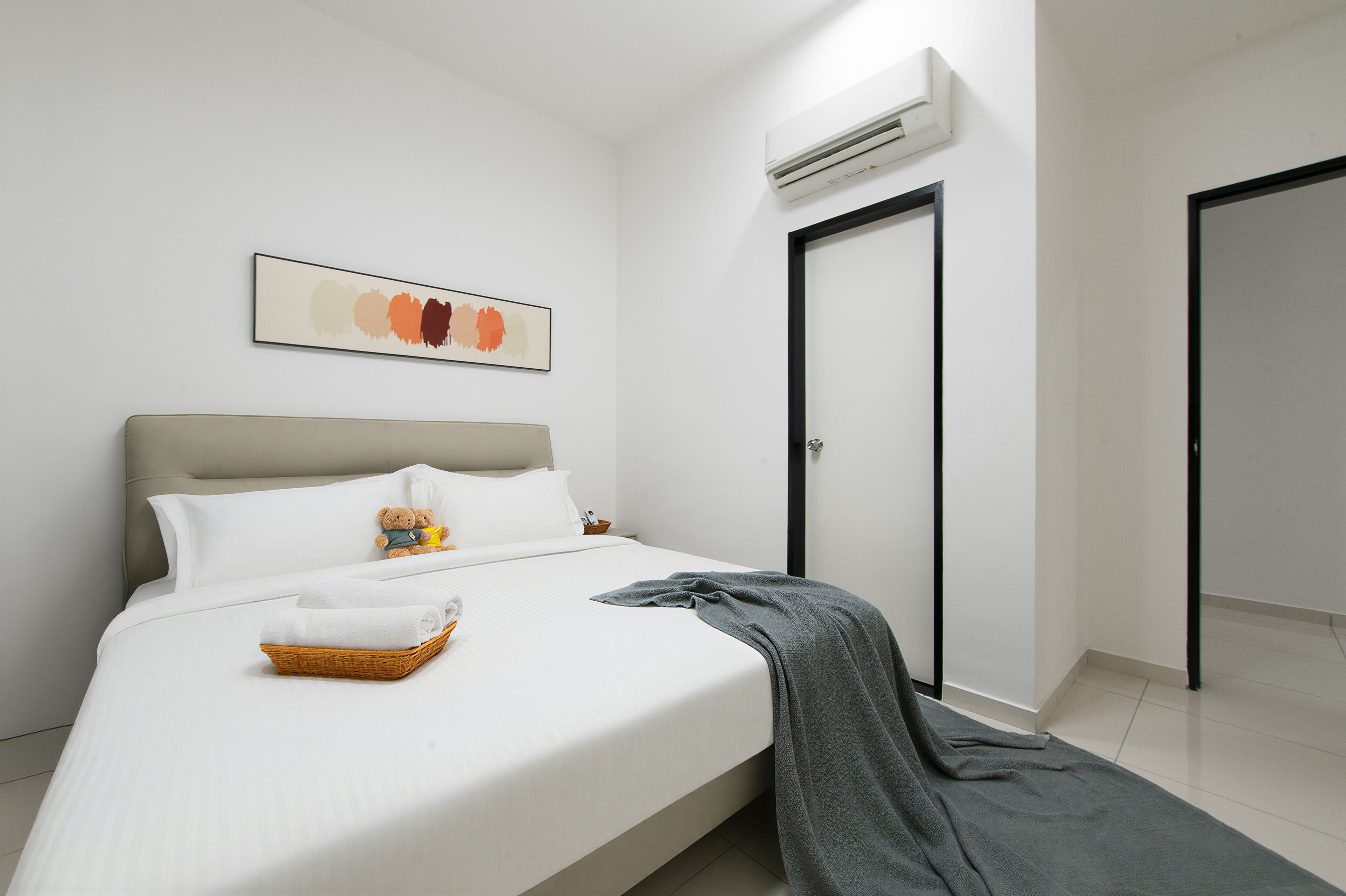 Una cama bien hecha con sábanas blancas, un plaid gris y una cesta de toallas dobladas. Hay un osito de peluche en la cama y un cuadro abstracto enmarcado en la pared.