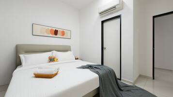 Una cama bien hecha con sábanas blancas, un plaid gris y una cesta de toallas dobladas. Hay un osito de peluche en la cama y un cuadro abstracto enmarcado en la pared.