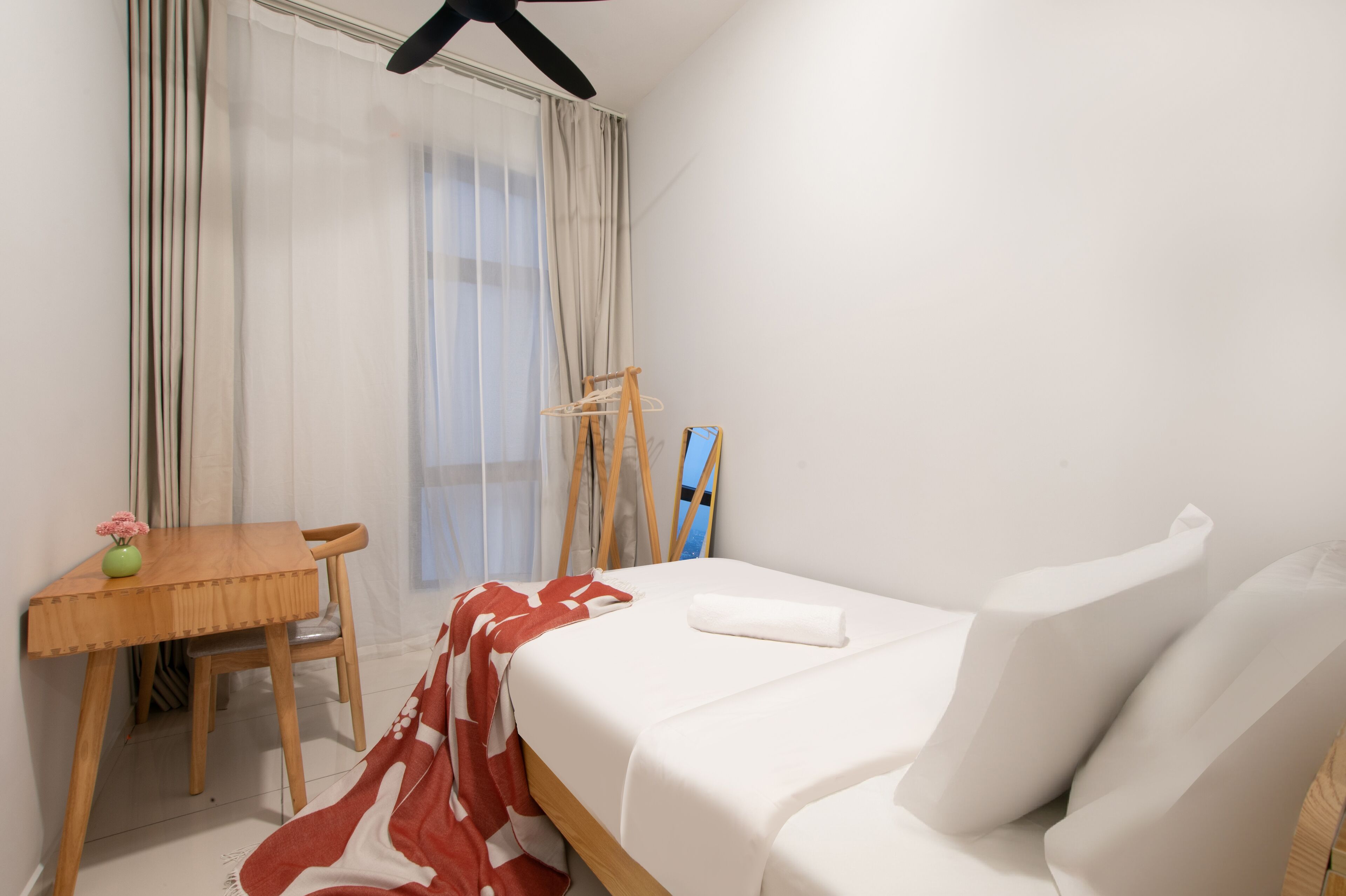 A hotel room with a bed, a wooden desk, a chair, a small table with a vase, and a window with curtains.
