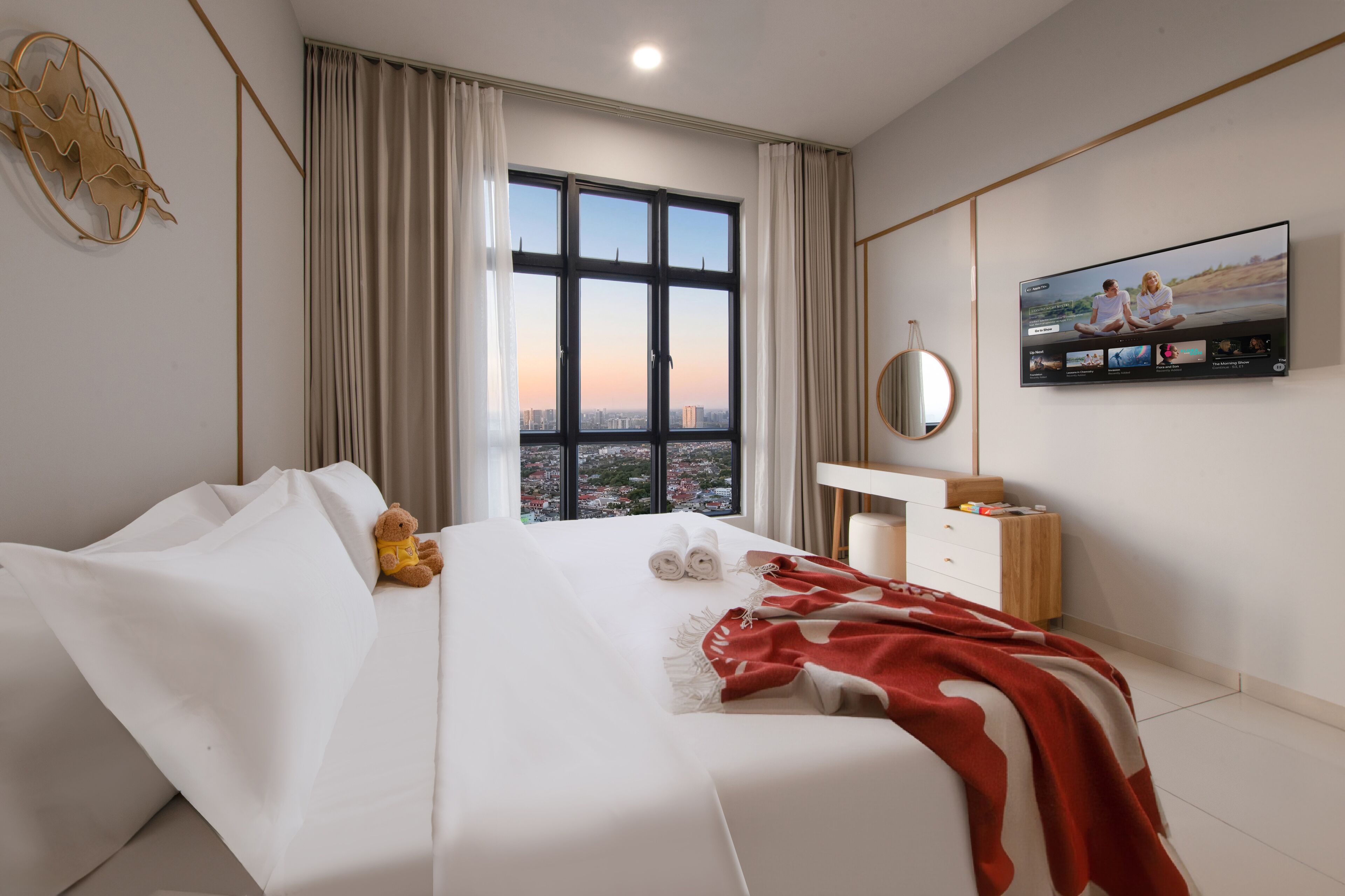 A hotel room with a large bed, a bedside table, a TV, and a window with a city view.