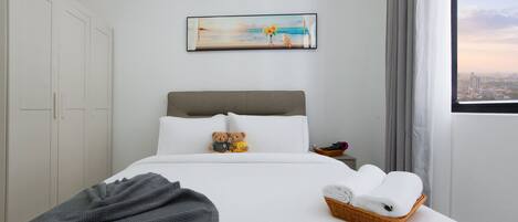 A hotel room with a bed, a painting of a beach scene, a window with a city view, and a basket of towels.
