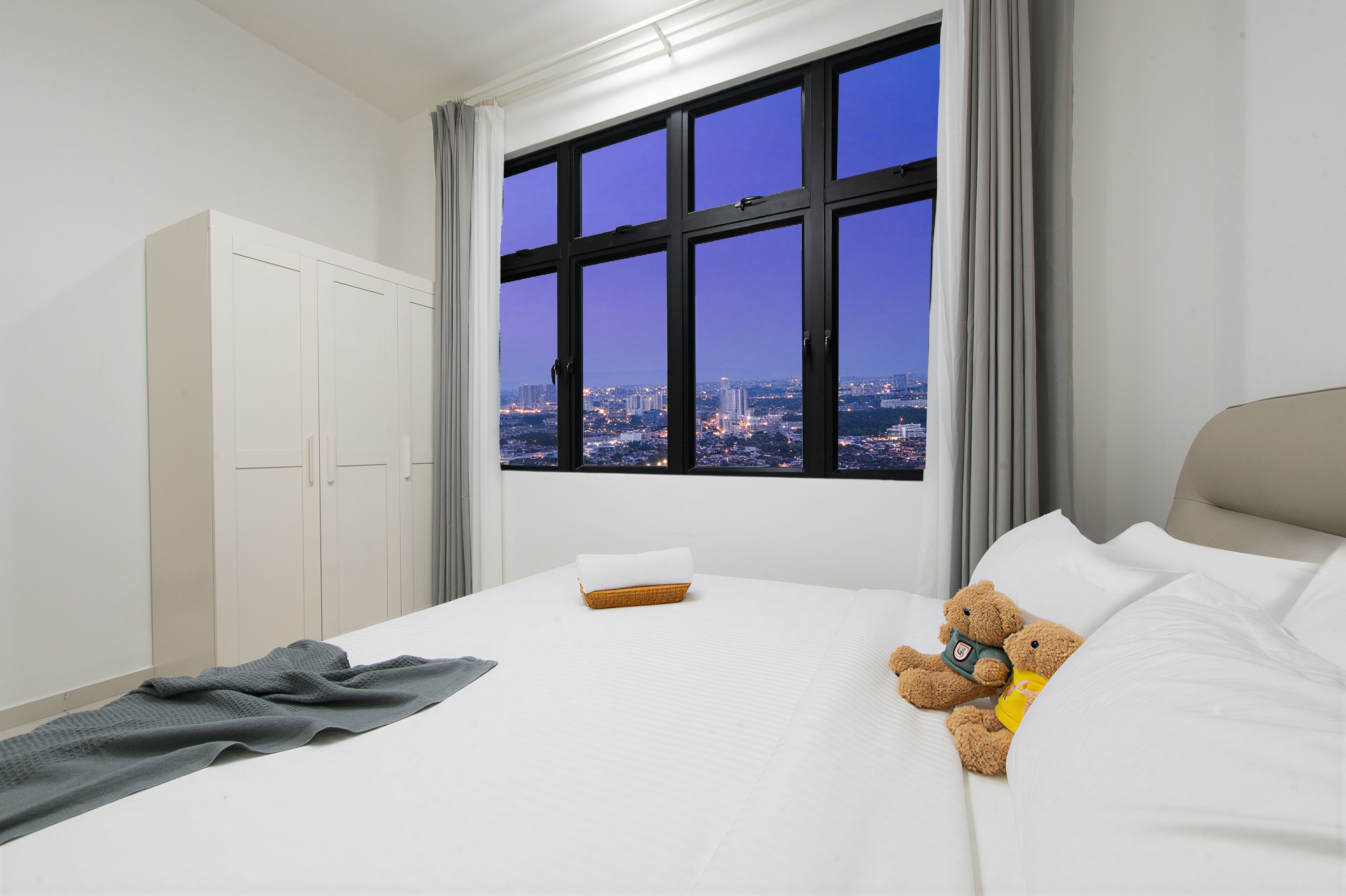 A hotel room with a large window, a bed with white linens, a teddy bear, and a grey blanket.