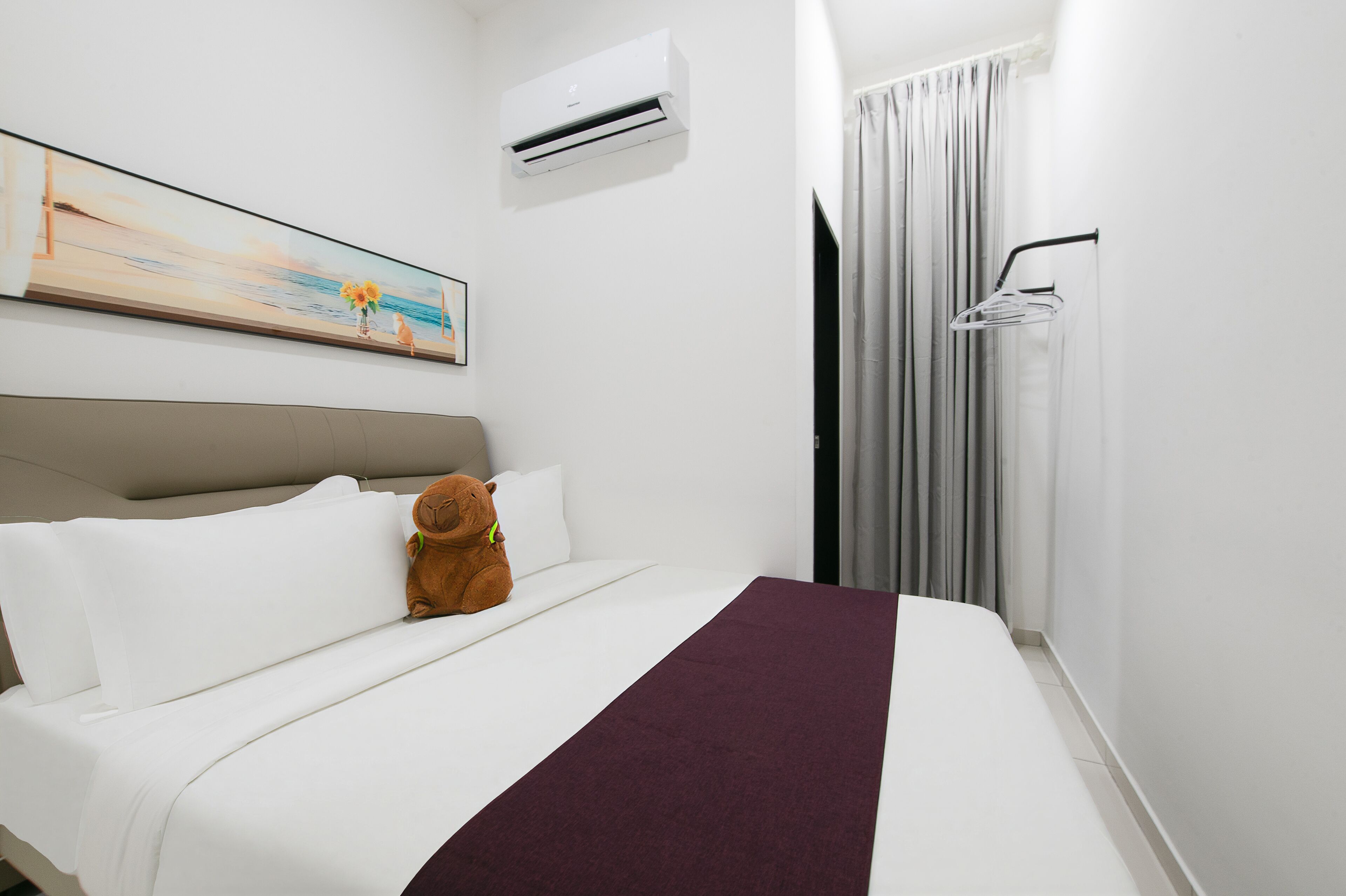 A hotel room with a bed, a teddy bear, a painting, an air conditioner, and a shower area.