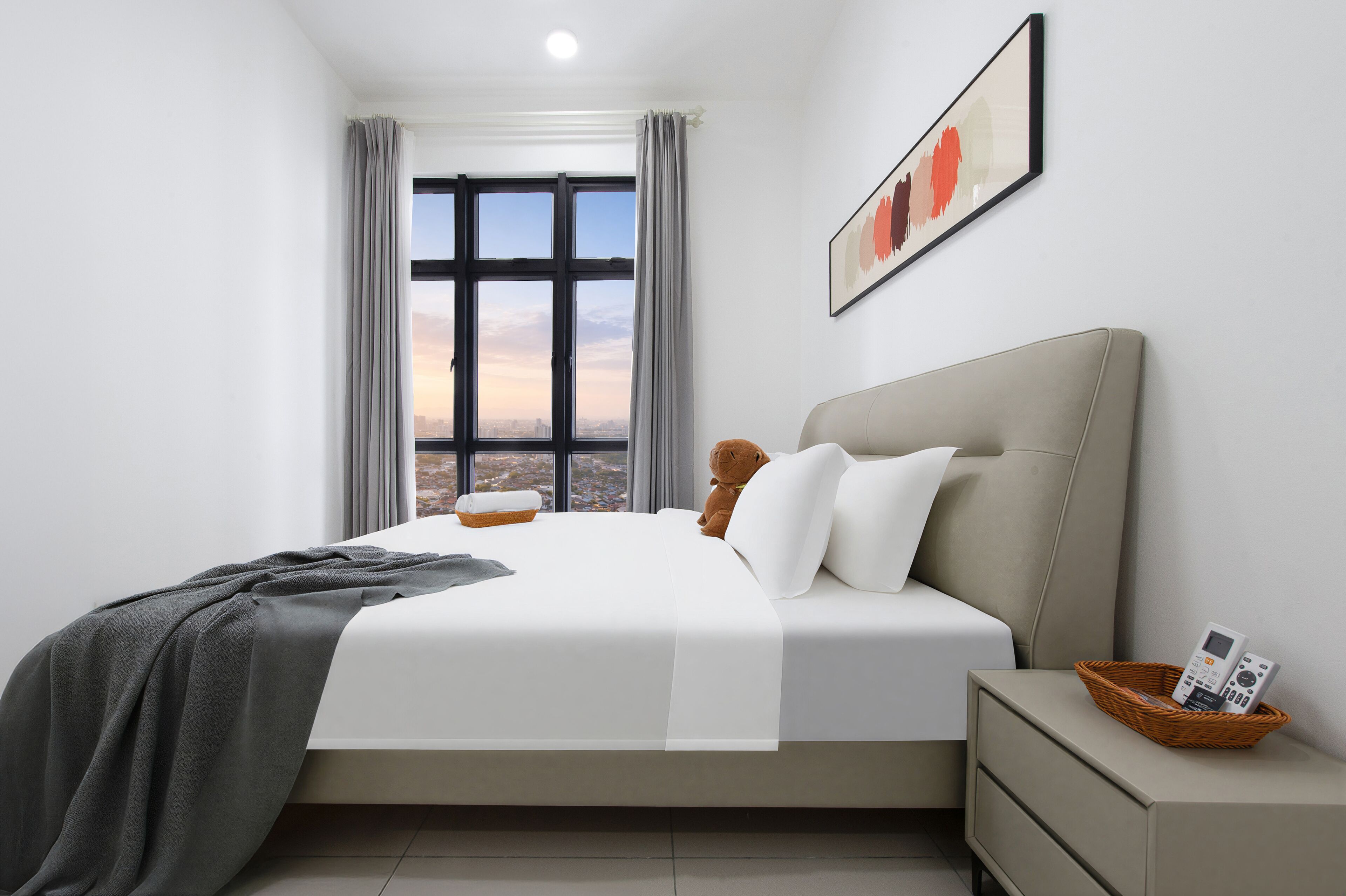 Une chambre d’hôtel moderne avec une grande fenêtre, un lit recouvert de draps blancs, un plaid gris, un ours en peluche, une table de chevet avec un panier, et un tableau abstrait encadré accroché au mur.