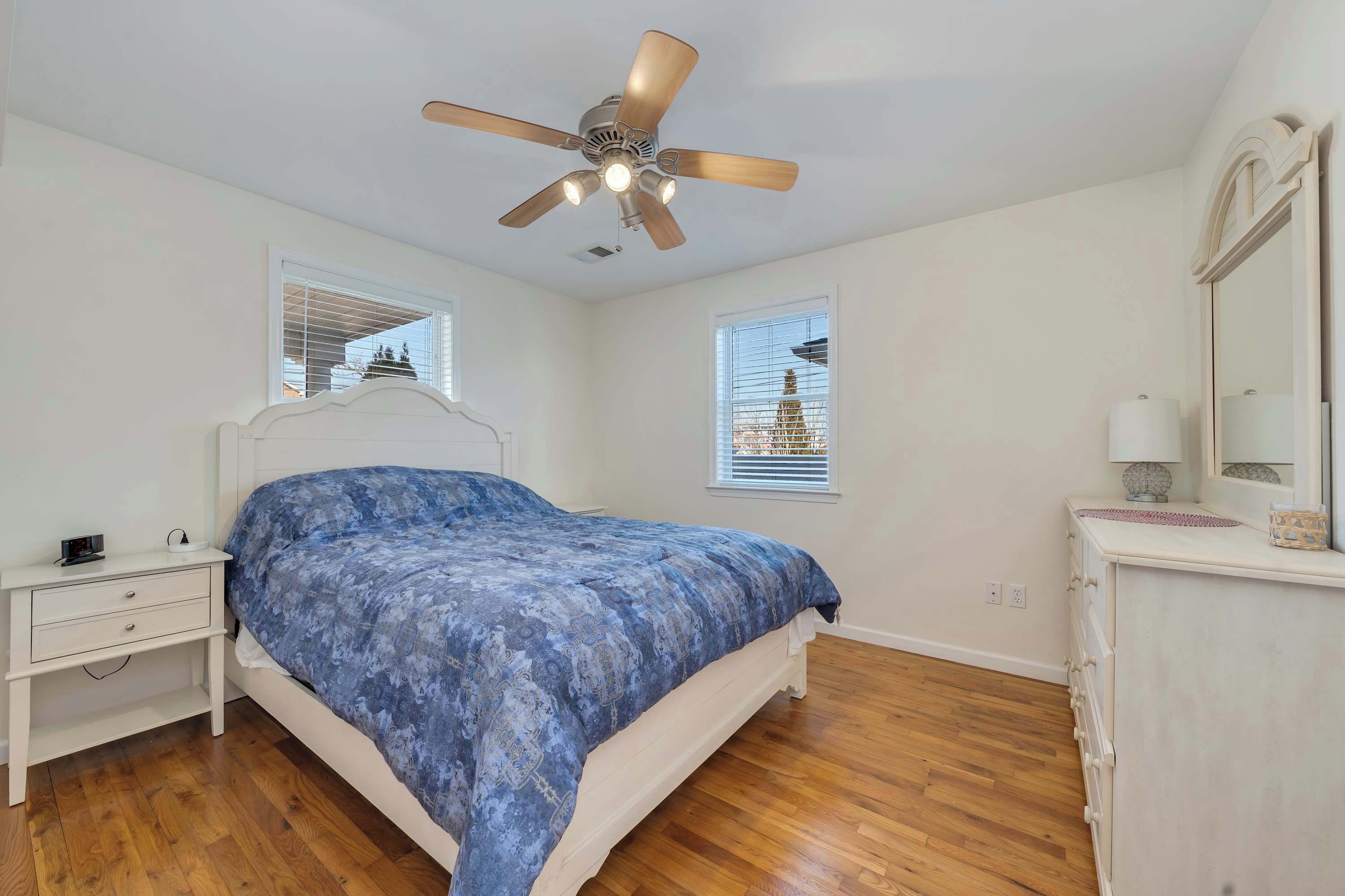 A bedroom with a bed, a nightstand, a dresser, a ceiling fan, and two windows with blinds.