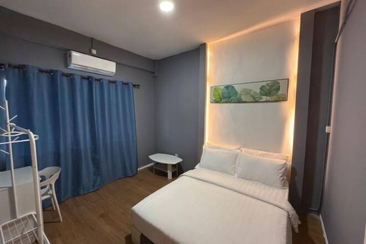 A bedroom with a bed, a small table, a chair, a wall-mounted air conditioner, and a painting.