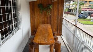 Une table en bois, entourée de six tabourets, se trouve sur un balcon doté d’une balustrade blanche et offrant une vue sur la rue.