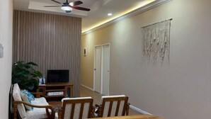 Un coin repas avec une table et des chaises en bois, un ventilateur de plafond, un téléviseur posé sur un meuble et un mur orné d’une œuvre en macramé.