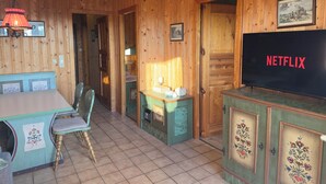A wooden cabin interior with a television displaying 'NETFLIX', a dining table, chairs, and a small kitchen area.