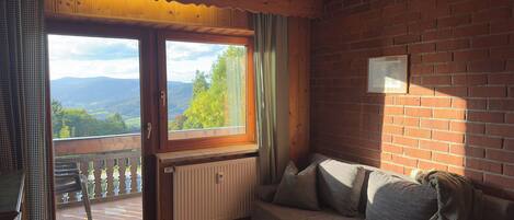 A cozy room with a wooden ceiling, a brick wall, a sofa with cushions, a radiator, and a window with a view of greenery and mountains.