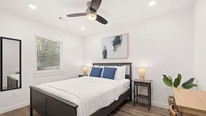 A bedroom with a bed, bedside tables, a ceiling fan, a window with blinds, a wall-mounted artwork, and a mirror.