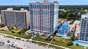 A high-rise hotel with multiple floors, a swimming pool, and a beach area with umbrellas.