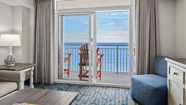 A hotel room with a large sliding glass door leading to a balcony with a view of the sea. There is a blue armchair and a small table with a lamp.