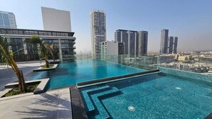 A rooftop swimming pool with clear blue water, surrounded by modern buildings and palm trees.