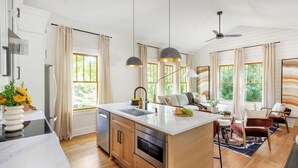 Une cuisine moderne dotée d’un îlot central, de meubles en bois et d’un four encastré. On y trouve également un coin repas avec un canapé et des chaises, ainsi que de grandes fenêtres qui laissent entrer la lumière naturelle.
