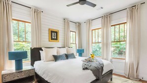 Une chambre dotée d’un grand lit, de deux tables de chevet, d’un ventilateur de plafond et offrant une vue sur la verdure par les fenêtres.