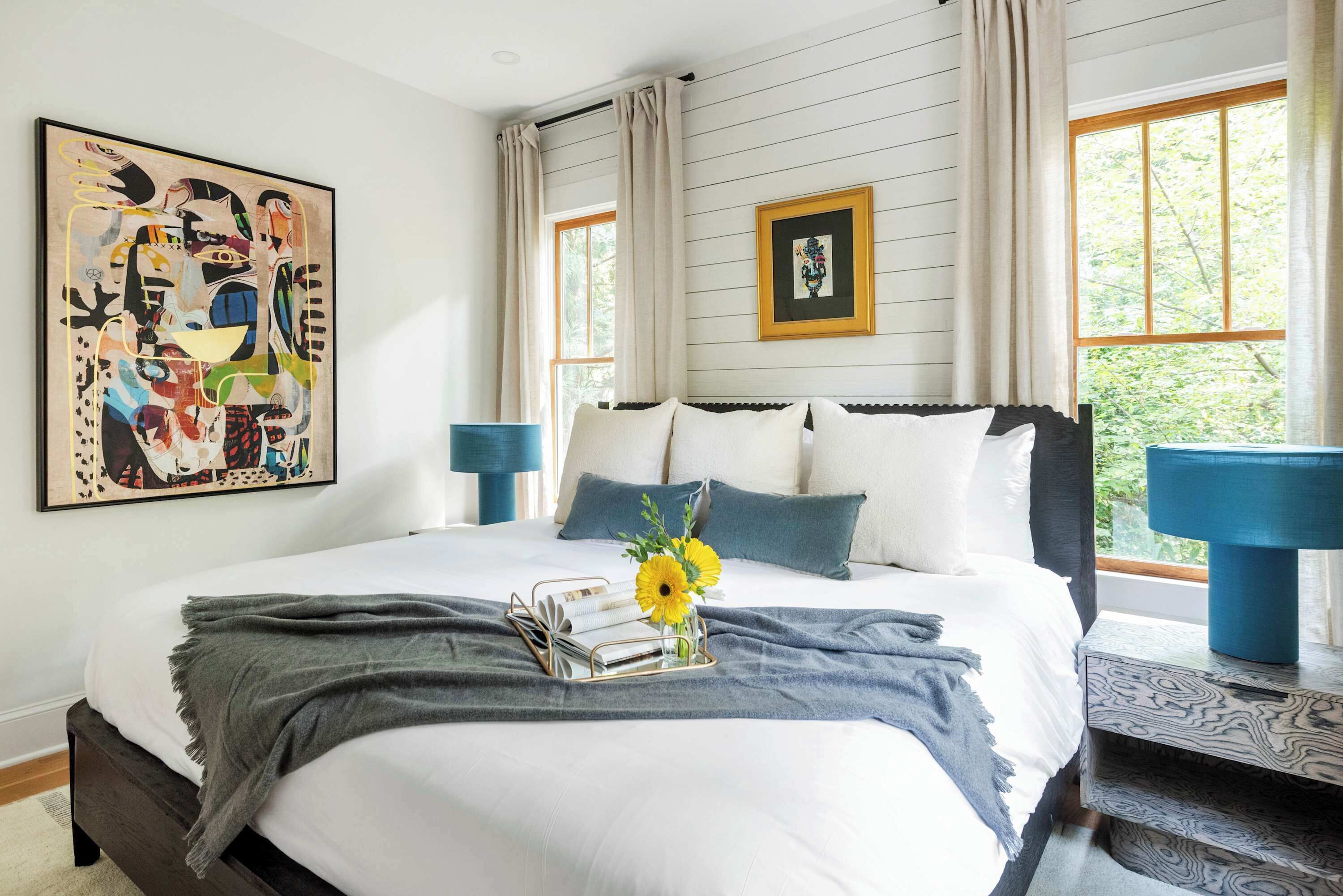 Un lit bien fait, recouvert d’un plaid gris, une table de chevet avec une lampe bleue et un grand tableau abstrait accroché au mur.