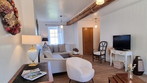 A cozy living room with a sofa, armchair, and a television. There is a wooden table with magazines, a lamp, and a wicker chair. A decorative wreath hangs on the wall, and a wooden ceiling beam is visible. - Charming and Comfortable Cottage in Saint-Haon-le-Châtel, Pets Allowed (Saint-Haon-le-Châtel)