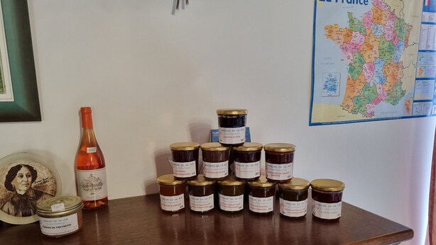 Une table en bois, sur laquelle on trouve plusieurs pots de confiture et une bouteille de vin, ainsi qu’un tableau encadré et une carte de la France accrochée au mur.