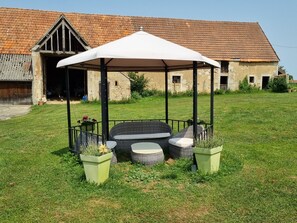 Un kiosque agrémenté de meubles en osier et de jardinières, situé dans un espace verdoyant devant un bâtiment en pierre surmonté d’un toit en tuiles.