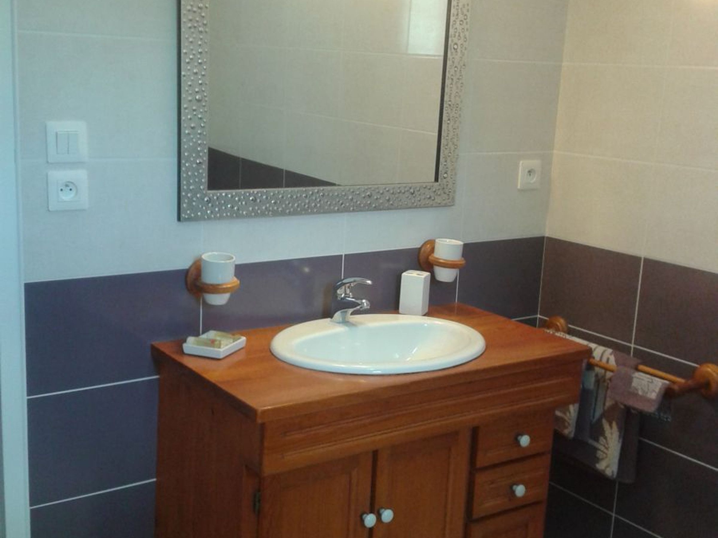 Une salle de bain avec un meuble-lavabo en bois, un lavabo blanc, un miroir avec un cadre décoratif et deux porte-gobelets.