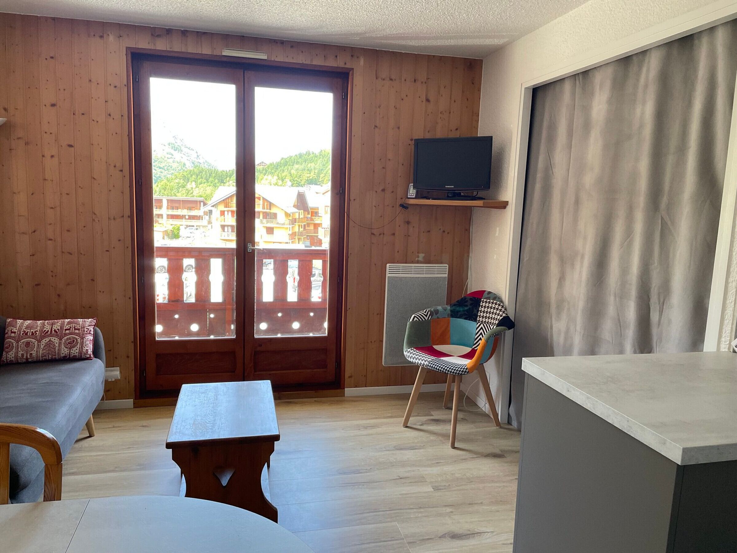 Ein Zimmer mit Holzwänden, einem Balkon mit Blick auf Gebäude, einer Couch, einem Tisch, einem Stuhl und einem Fernseher auf einem Regal.