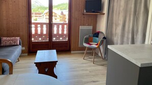 A room with wooden walls, a balcony with a view of buildings, a sofa, a table, a chair, and a TV on a shelf. - Studio cabin for 4 people, 3* with NW facing balcony (Valloire)