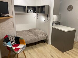 Ein kleines Zimmer mit einem Bett, einem kleinen Schreibtisch, einem Sessel, einem Fernseher und einem Projektor.