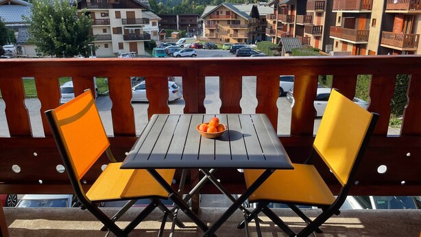 A balcony with two yellow chairs and a table, overlooking a parking area and buildings with wooden balconies, set against a mountainous backdrop. - Studio cabin for 4 people, 3* with NW facing balcony (Valloire)