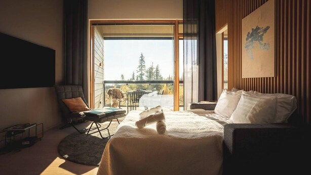 A modern hotel room with a large bed, a seating area with a chair and a small table, a balcony with a view of trees, and a painting on the wall.