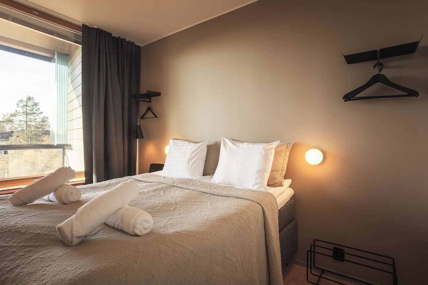 Ein ordentlich bezogenes Bett mit aufgerollten Handtüchern, einer Nachttischlampe und Blick ins Freie durch ein Fenster.