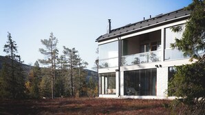 A modern two-story house with large windows and a balcony, surrounded by trees and a hilly landscape.