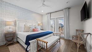Un dormitorio con cama, lámparas de noche, una silla de madera, televisor y un balcón con cortinas.