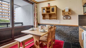 Een eetgedeelte met een houten tafel en stoelen, een rood tapijt en een stenen muur. Er is een raam met uitzicht op gebouwen.