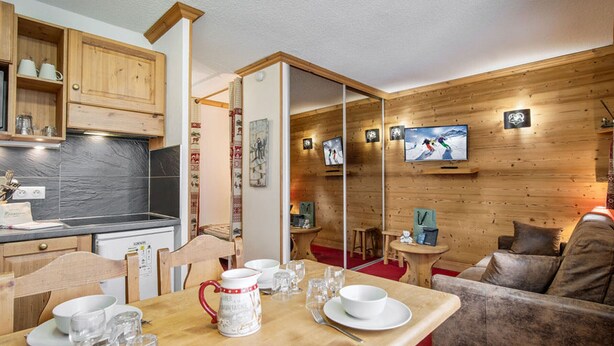 Een gezellige kamer met een houten eettafel voor vier personen, een bank, een aan de muur bevestigde televisie, en een keuken met houten keukenkastjes.