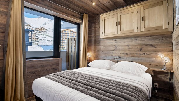 A bedroom with a wooden ceiling, a large bed with white and grey bedding, a window with curtains, and a wooden cabinet above the bed.