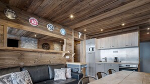 A cozy cabin interior with a wooden ceiling, a sofa with patterned cushions, a dining table, and a kitchen area with cabinets and appliances.