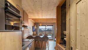 Een moderne keuken met houten kastjes, een eethoek met stoelen en uitzicht op bergen door een groot raam.