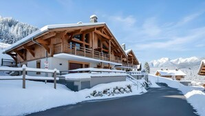 A wooden chalet with a balcony, surrounded by snow-covered mountains and a clear sky.