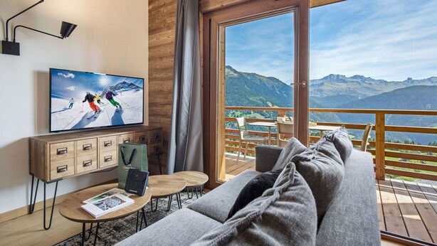A modern room with a wooden ceiling, a television displaying a ski scene, a sofa, a small table, and a view of mountains through a large sliding door.