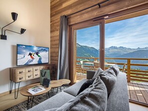 Habitació moderna amb sostre de fusta, televisió que mostra una escena d'esquí, un sofà, una taula petita i vista a les muntanyes a través d'una gran porta corredissa.