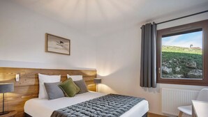 Ein Schlafzimmer mit einem hölzernen Kopfende, einem Bett mit grau-weißer Bettwäsche, einer grauen, gepolsterten Bank, einem Nachttisch mit Lampe, einem Sessel und einem Fenster mit Vorhängen.