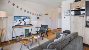 Ein modernes Wohnzimmer mit einem grauen Sofa, einem Holztisch, einem Fernseher, der einen Skifahrer zeigt, und einer Küchenzeile mit einer Mikrowelle und Holzschränken.