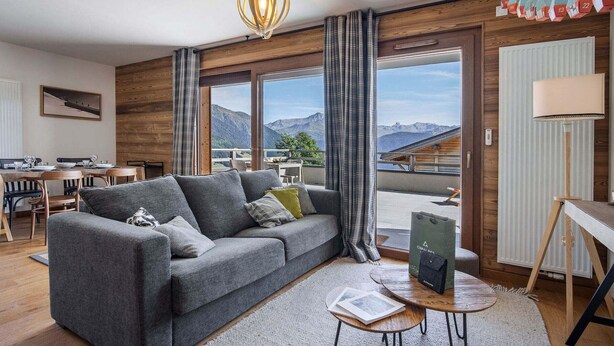 Ein modernes Wohnzimmer mit grauem Sofa, Holzakzenten, einem Essbereich mit Tisch und Stühlen und Blick auf Berge durch große Fenster.
