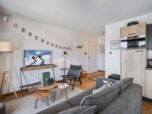 A modern living room with a grey sofa, a wooden coffee table, a TV displaying a skier, and a kitchen area with a microwave and wooden cabinets.
