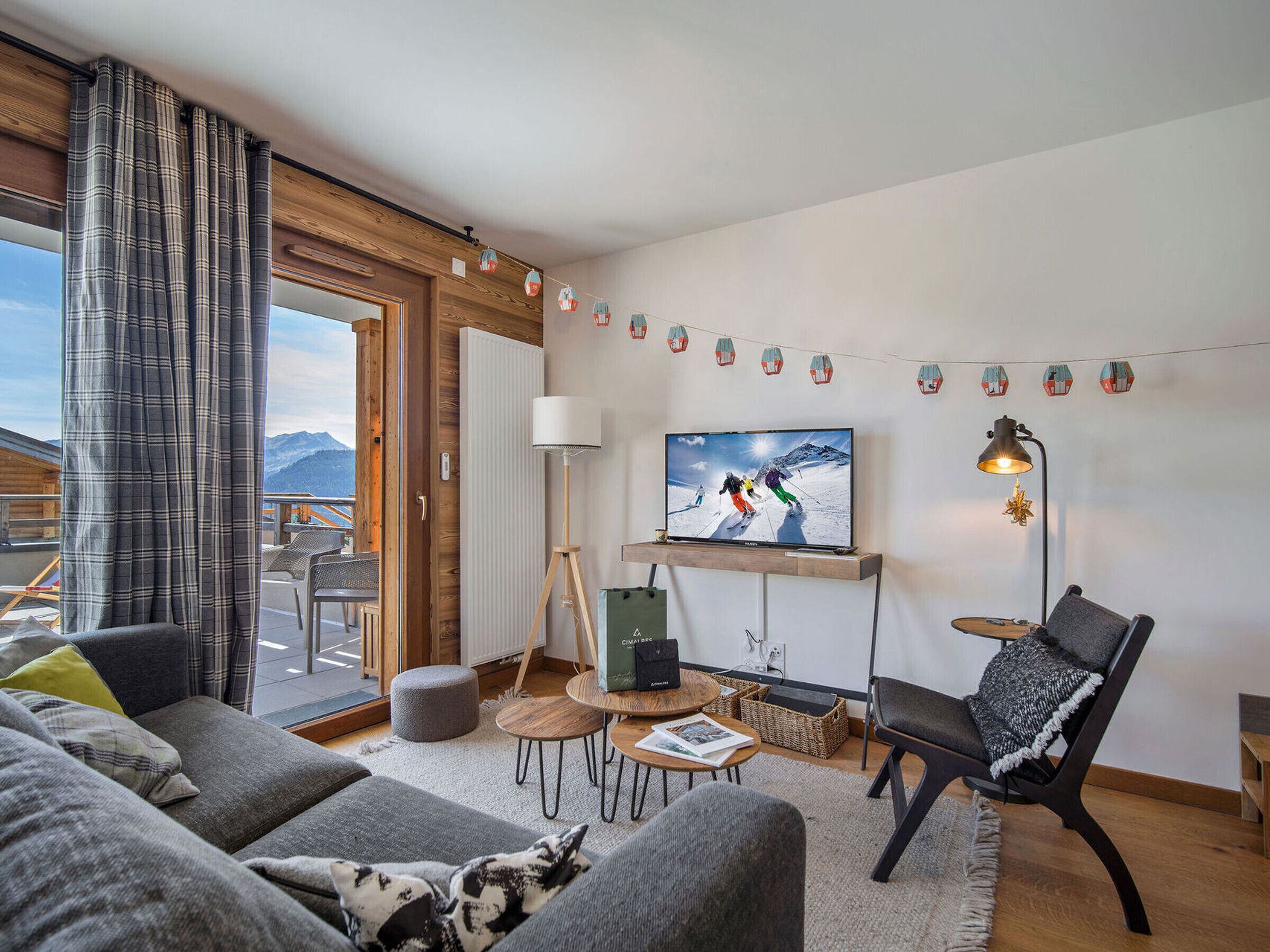A modern living room with a large TV, comfortable seating, a wooden floor, and a view of mountains through the window.