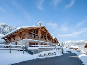 A wooden chalet with a balcony, surrounded by snow-covered mountains and a clear sky.
